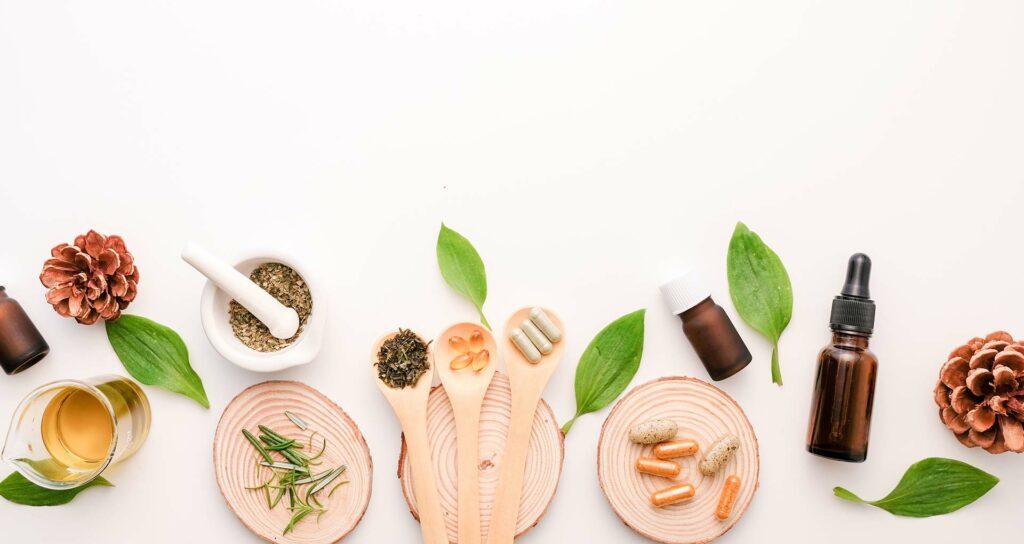 Mortar and pestle, spoons of herbs and capsules, and amber dropper bottles on a white surface