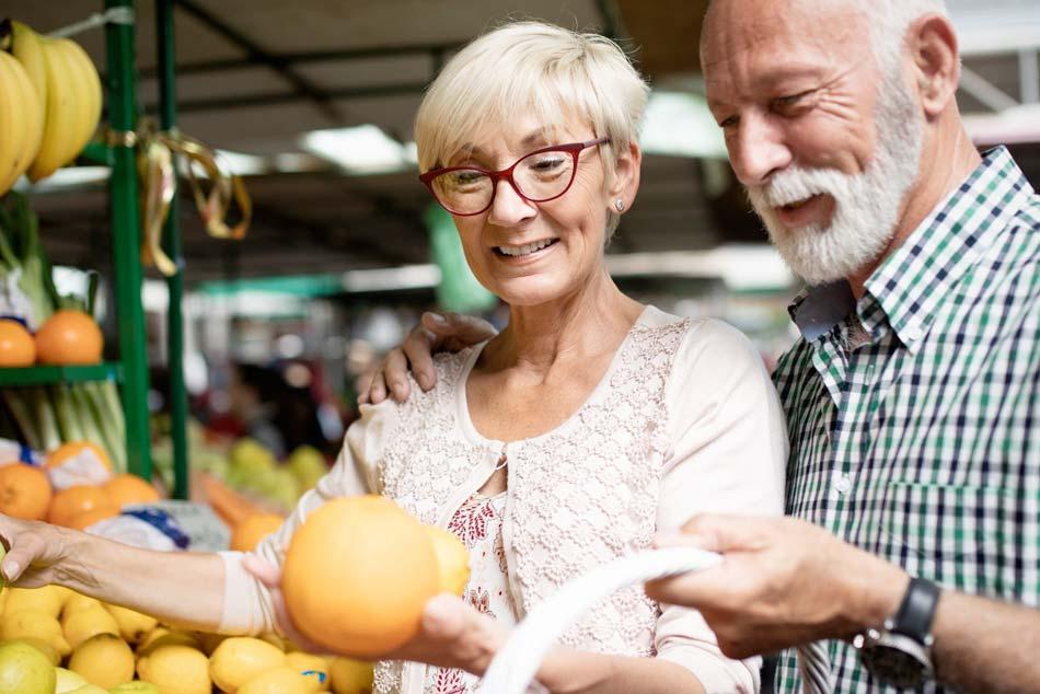 An older woman in red glasses holding up an orange beside a gray-bearded man at a produce market