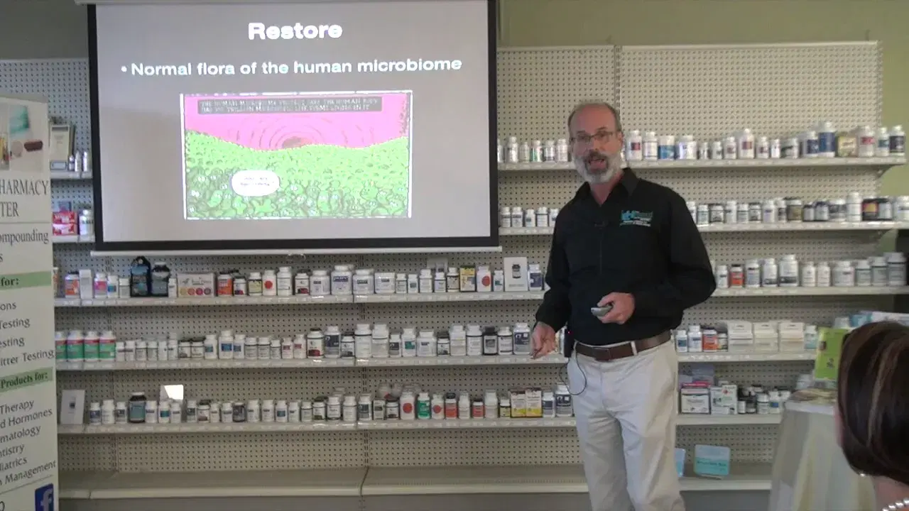 A presenter beside a projected slide titled Restore, in front of shelves of supplement bottles