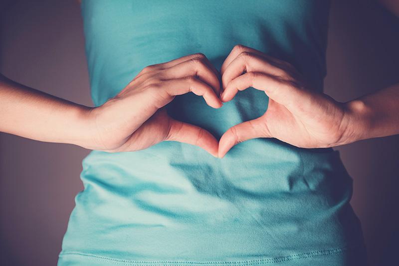 Hands forming a heart shape over the stomach of a person in a teal top