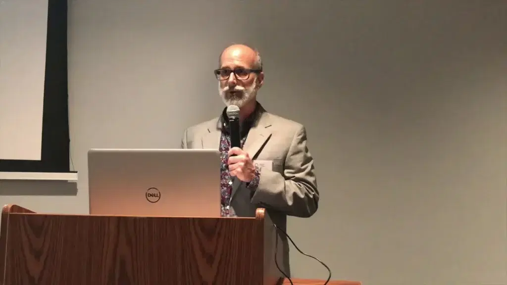 A bearded speaker in glasses and a tan blazer at a podium microphone