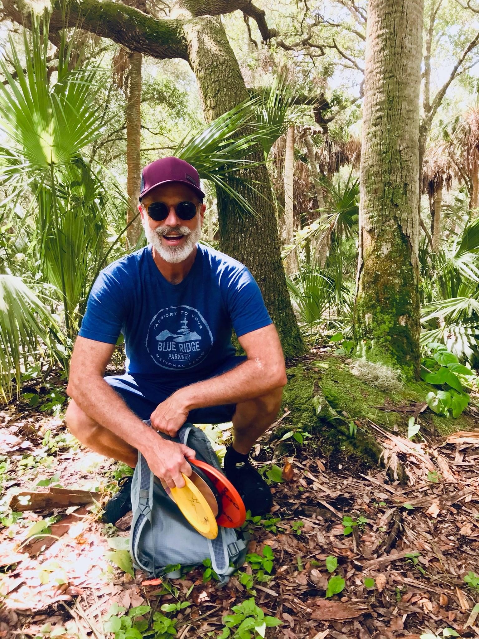 A man in a cap and sunglasses crouching in a palm forest with a backpack and flying discs