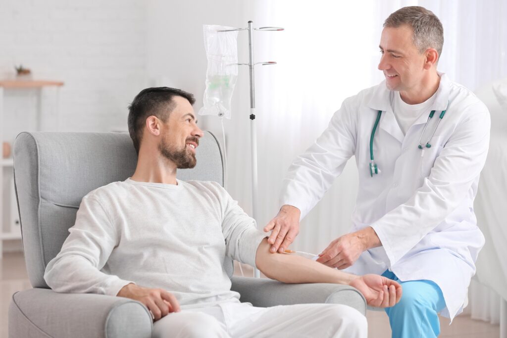 A smiling man in an armchair as a clinician in a white coat attends to his arm beside an IV stand